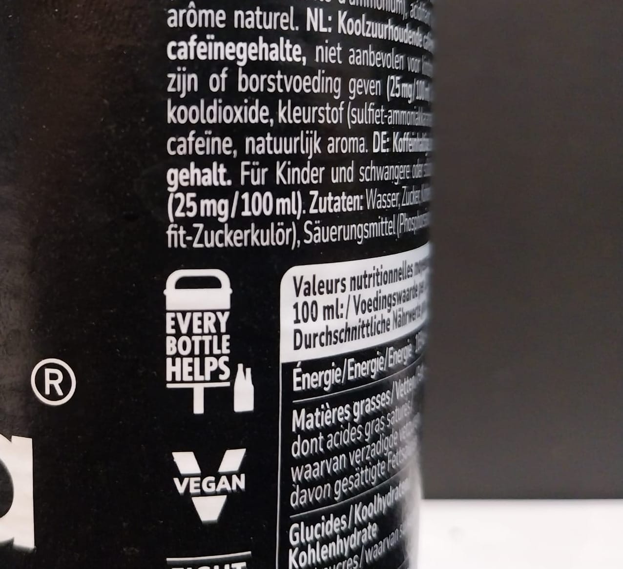 The side of a cola bottle, showing an incomplete view of ingredients and nutritional information, with a focus on an icon which shows a raised garbage can with bottles under it and the slogan "Every bottle helps."