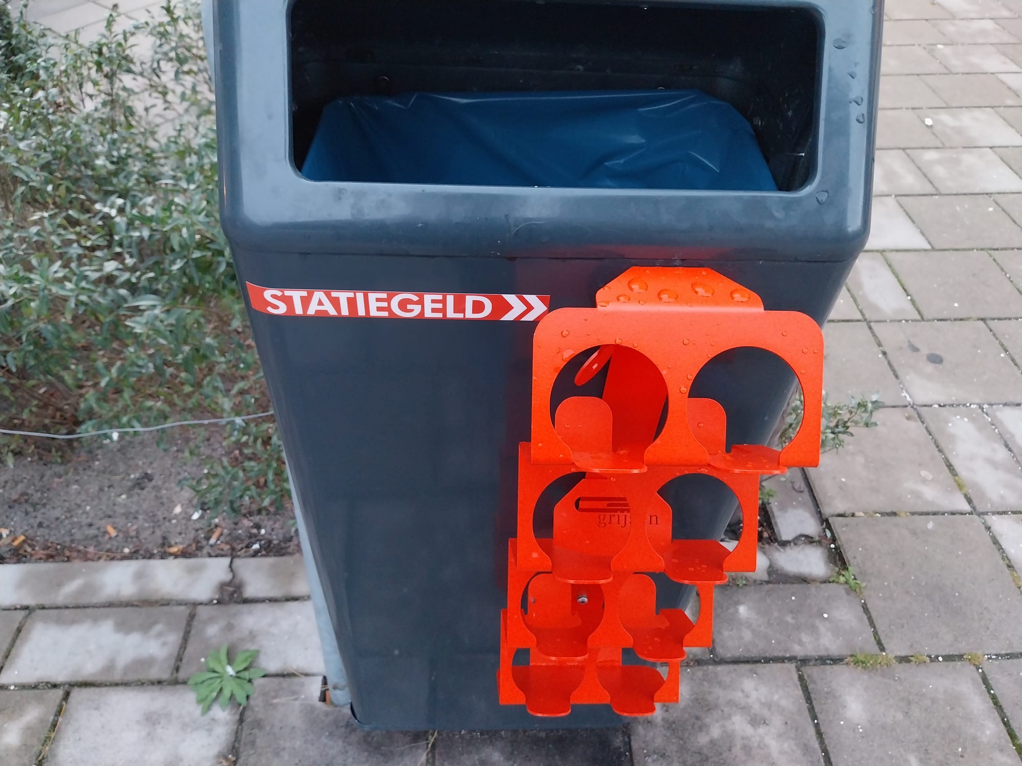Grey garbage bin on a sidewalk which has an orange rack attached to its side for bottle collection.