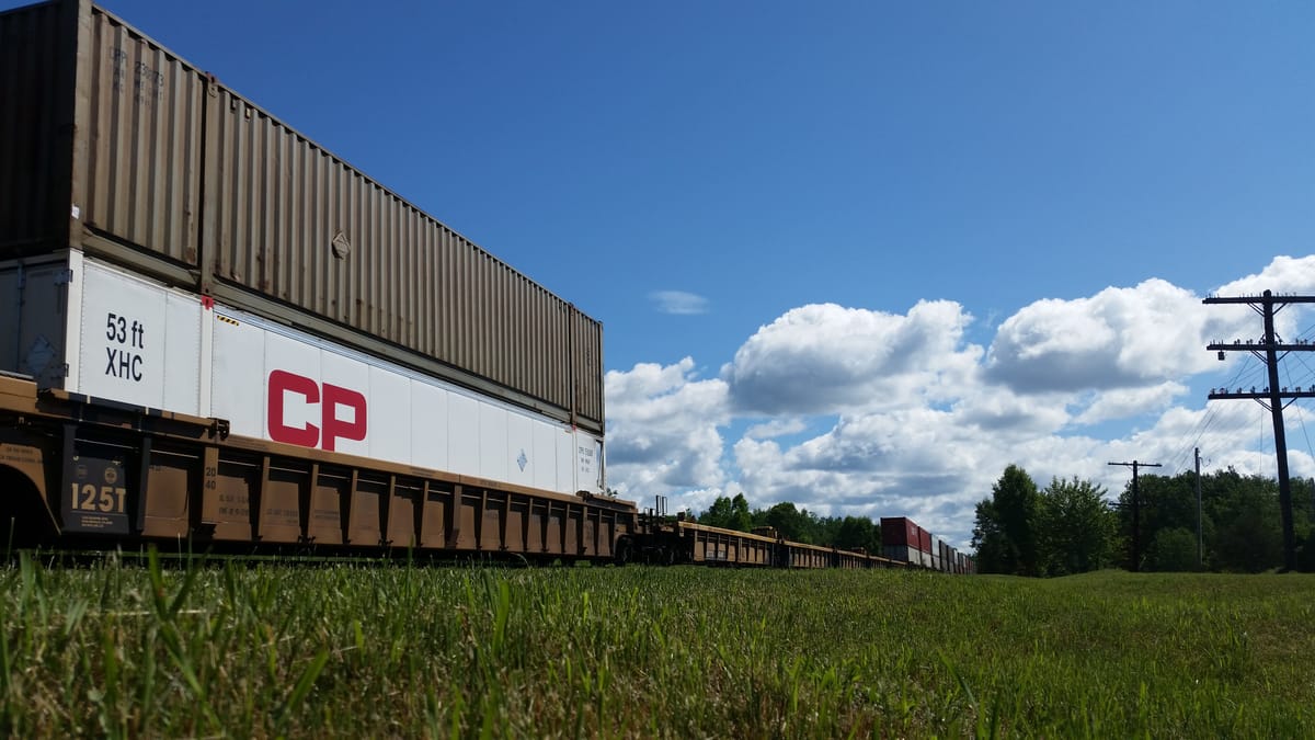 Photo of a train carrying intermodal shipping containers. In the foreground, a grassy field. In the background, a blue sky with fluffy clouds.