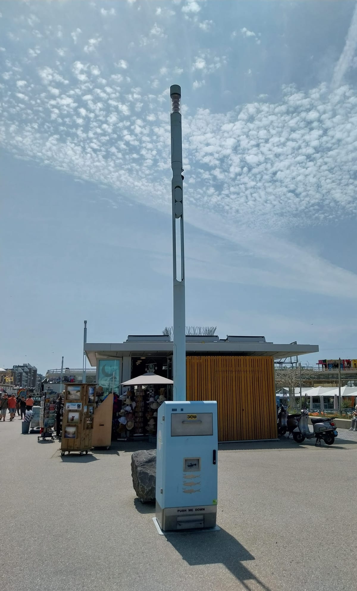 Photo with a garbage bin and light post, both light blue. The sky is a similar shade of blue, and there is a shop in the background selling hats and souvenirs. 
