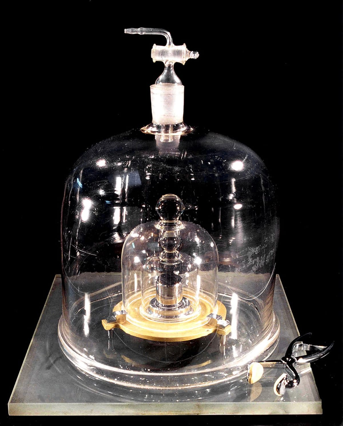 A photo of a small, metal weight within several layers of protective glass cloches. A pair of tongs are placed next to the outermost cloche. The background of the photo is black.