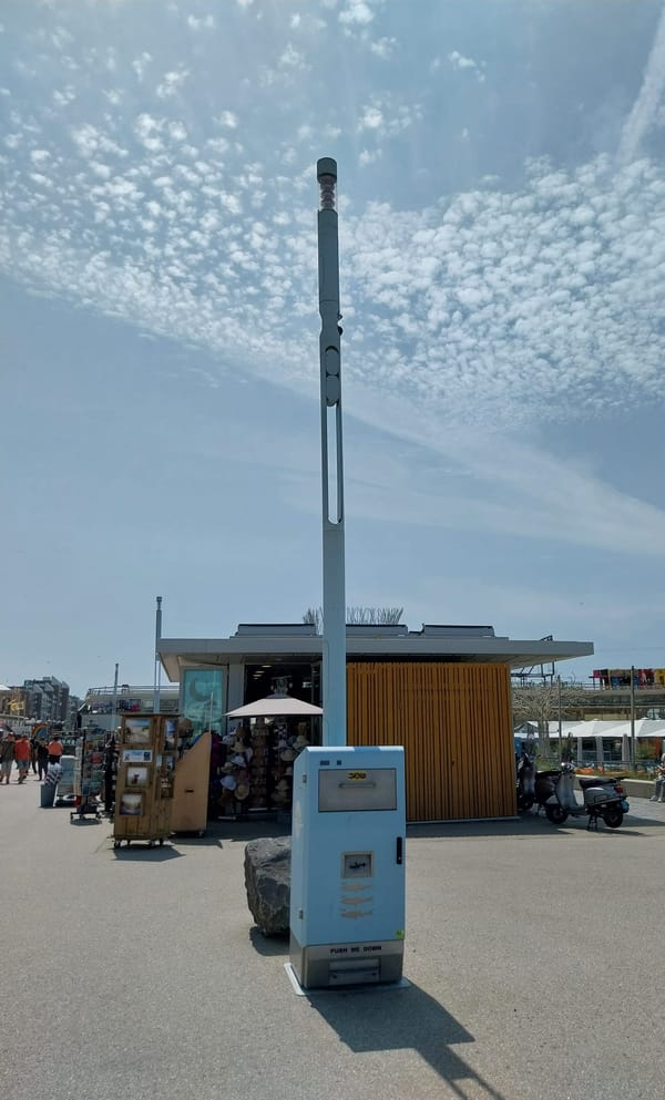 Photo with a garbage bin and light post, both light blue. The sky is a similar shade of blue, and there is a shop in the background selling hats and souvenirs. 