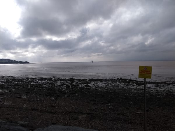 Photo of a beach and a bright but cloudy sky. There is a ship far away on the horizon, and a yellow sign in the foreground that says "Danger Soft Mud."