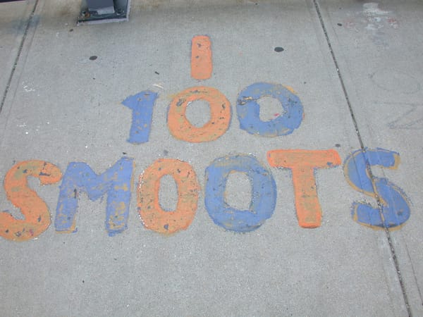 On a sidewalk, bubble writing in blue and red which reads "100 SMOOTS."